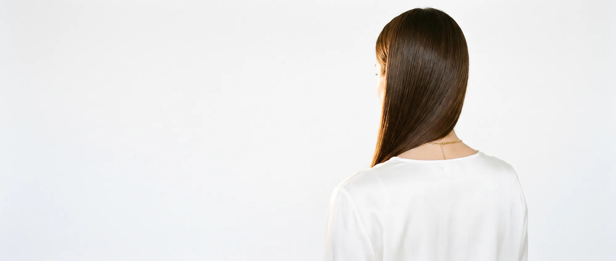 Una donna di spalle con lunghi capelli castani lisci e lucidi, su sfondo bianco