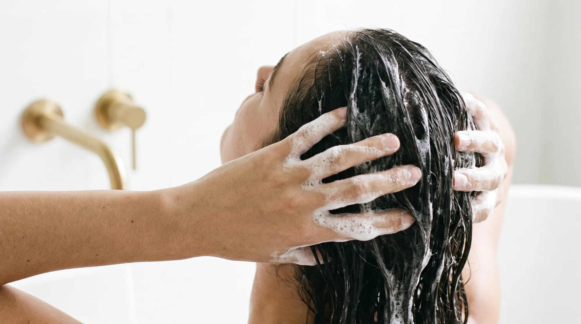 Due mani massaggiano la schiuma dello shampoo sui capelli scuri bagnati, in un bagno bianco con rubinetti dorati
